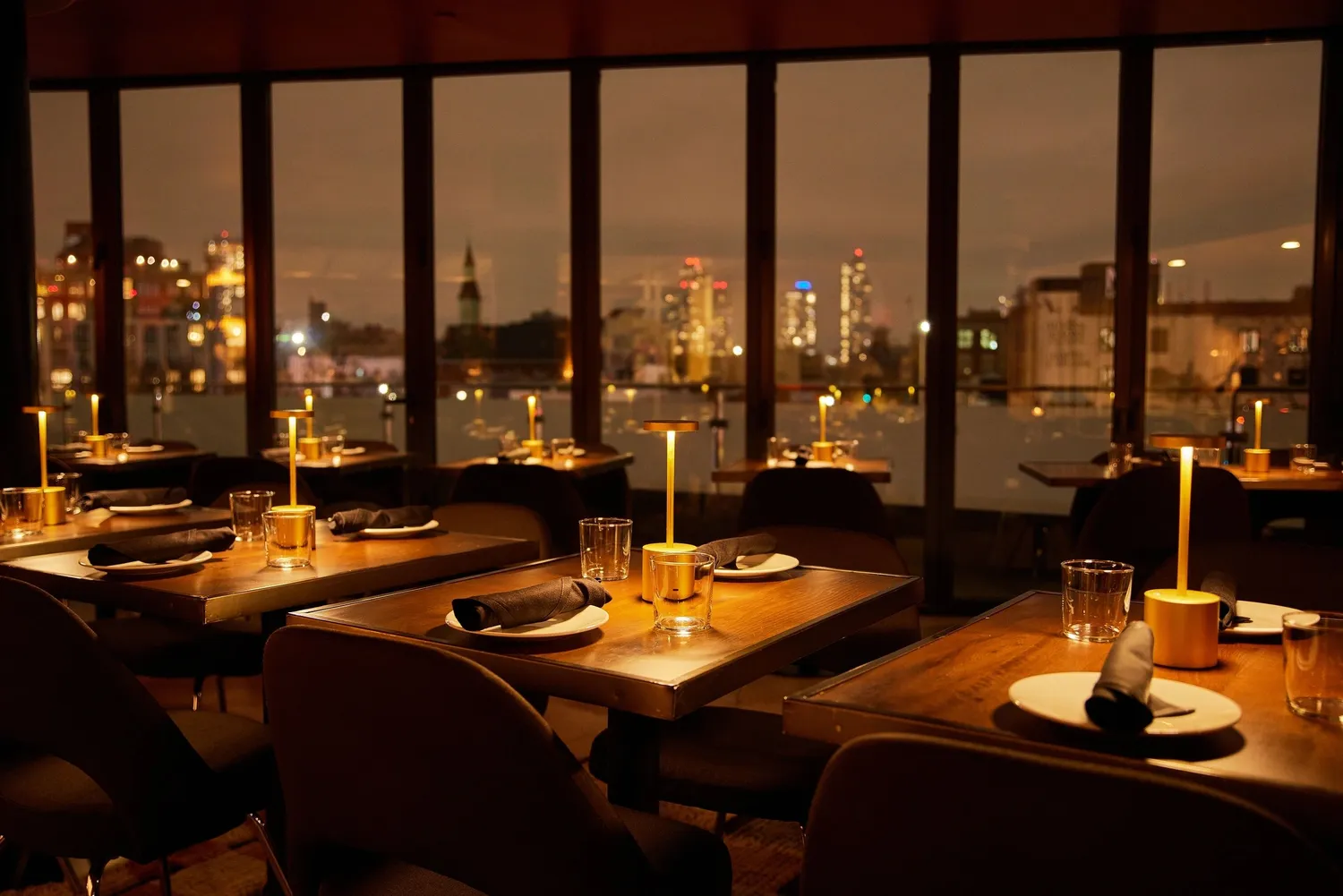 Cozy restaurant interior with wooden tables, elegant candles, and city skyline visible through large windows at night.