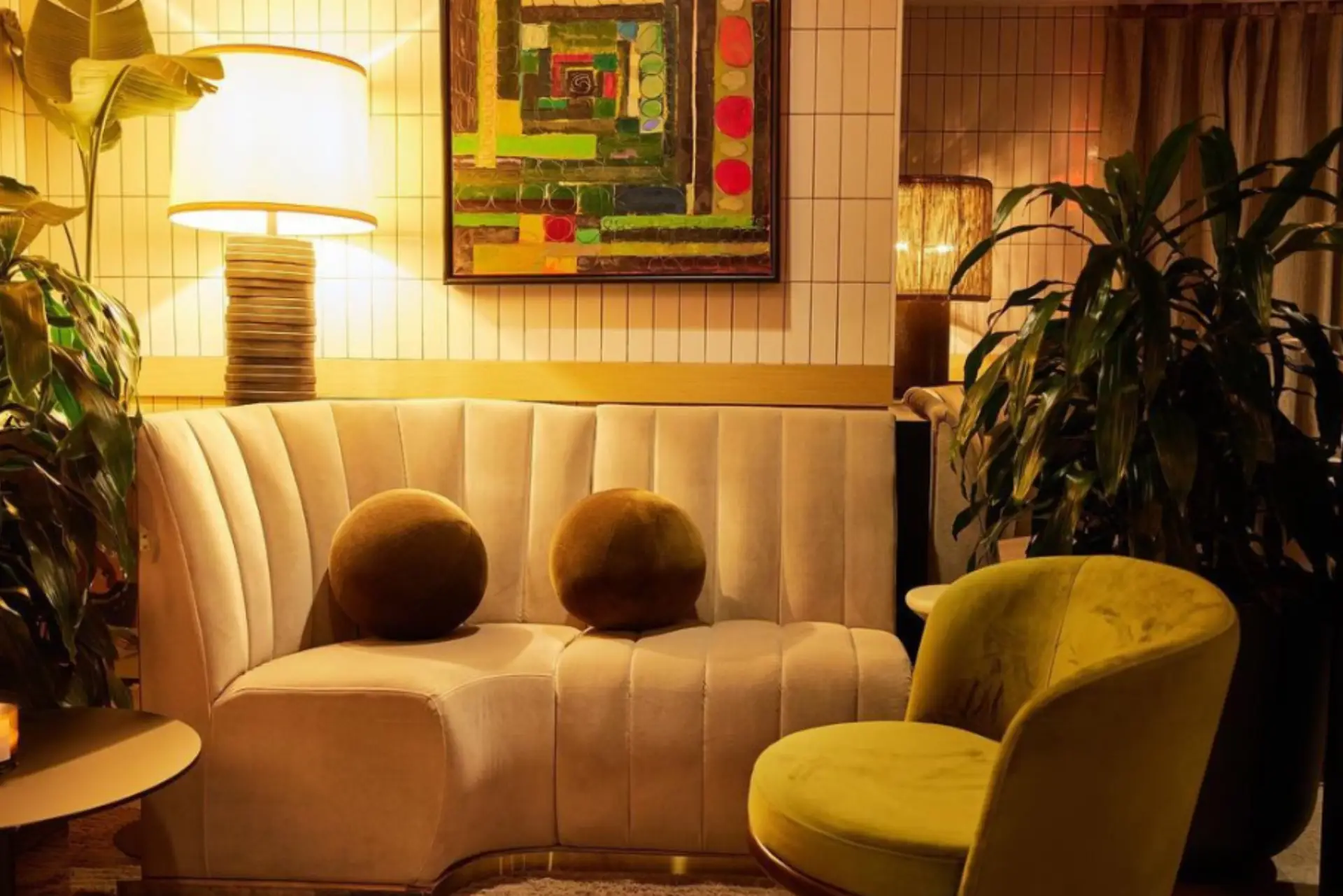 A cozy seating area featuring a curved beige sofa, round green cushions, a vibrant painting, and warm lighting from lamps.