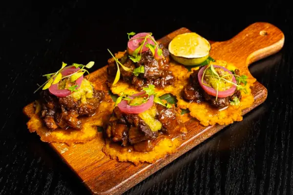 Four mini tacos topped with savory beef, pickled onions, and green salsa, served on a wooden board with lime wedges and fresh herbs.